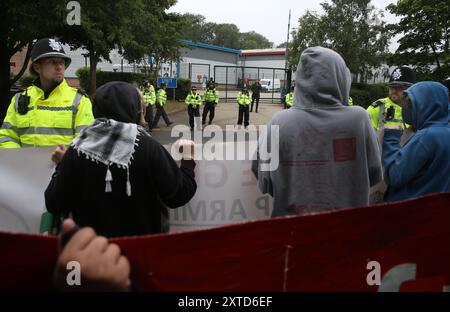 Leicester, Leicestershire, Regno Unito. 14 agosto 2024. Gli agenti di polizia stanno tra i manifestanti e il cancello della fabbrica della Elbit Systems durante la dimostrazione. I manifestanti si riuniscono fuori dai cancelli della fabbrica Elbit System a Leicester. Gli operatori sanitari per la Palestina si sono Uniti alla manifestazione molti di loro nei loro scrub per protestare mentre i lavoratori venivano a lavorare presso la compagnia israeliana di armi. I manifestanti protestano contro le armi fabbricate in questa fabbrica che ritengono vengano successivamente utilizzate per opprimere i palestinesi a Gaza e altrove. Il 85% della flotta di droni israeliani è fornito da Elbit Systems A. Foto Stock