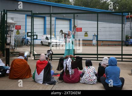 Leicester, Leicestershire, Regno Unito. 14 agosto 2024. I manifestanti si siedono sul pavimento di fronte ai cancelli durante la dimostrazione indirizzata da uno dei loro colleghi. I manifestanti si riuniscono fuori dai cancelli della fabbrica Elbit System a Leicester. Gli operatori sanitari per la Palestina si sono Uniti alla manifestazione molti di loro nei loro scrub per protestare mentre i lavoratori venivano a lavorare presso la compagnia israeliana di armi. I manifestanti protestano contro le armi fabbricate in questa fabbrica che ritengono vengano successivamente utilizzate per opprimere i palestinesi a Gaza e altrove. Il 85% della flotta di droni israeliani è fornito da Elbit System Foto Stock