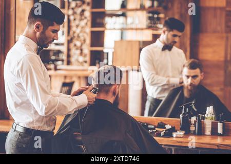 Rifilatura del nastro. Vista posteriore del giovane barbuto che viene tagliato dal parrucchiere con rasoio elettrico mentre si siede sulla sedia del barbiere di fronte allo specchio Foto Stock