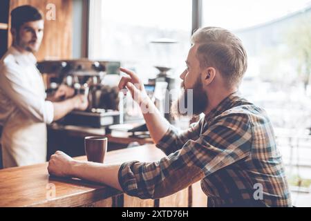 Un altro caffè, per favore! Giovane, bello e barbuto che parla con il barista mentre si siede al bancone del bar al bar Foto Stock