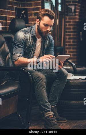 Controllo dell'elenco degli appuntamenti. Bel giovane uomo barbuto che lavora su un tablet digitale mentre si siede su una comoda sedia al barbiere Foto Stock