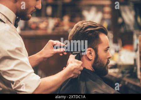 Styling professionale. Close up vista laterale del giovane uomo barbuto getting haircut dal parrucchiere con rasoio elettrico al barbiere Foto Stock