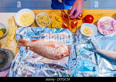 Guardando verso il basso come un uomo affetta i pomodori come parte della preparazione di un pasto con dentice rosso, verdure e altri recipienti di ingredienti tritati. Foto Stock