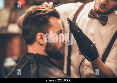 Già bello. Primo piano vista laterale di giovane uomo bearded che si cura dal parrucchiere con asciugacapelli al barbiere Foto Stock