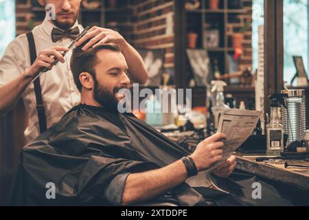 Siamo stati benissimo al barbiere. Allegro giovane uomo barbuto che viene tagliato da parrucchiere e legge il giornale mentre si siede sulla sedia al barbiere Foto Stock