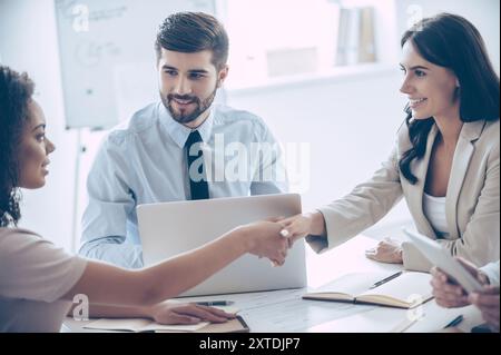 Piacere di conoscervi! Due belle donne che stringono la mano con il sorriso mentre si siedono al tavolo dell'ufficio con la loro collega Foto Stock