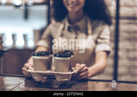 Il miglior caffè da portare via! Parte di una giovane donna africana allegra in grembiule che tiene tazze di caffè mentre si trova in piedi al bar Foto Stock