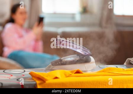 Primo piano del ferro da stiro a vapore sull'asse da stiro. In background, la donna sfocata è seduta a riposare sul divano e a usare lo smartphone. Concetto di famiglia fi Foto Stock