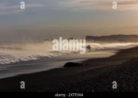 Le onde delle sneaker si infrangono sulla costa di Reynisfjara Black Sand Beach, con forti venti che soffiano acqua e sabbia nera vulcanica, tramonto, Islanda Foto Stock