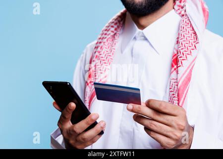 L'uomo arabo tiene le mani con carta di credito e smartphone mentre effettua l'acquisto in un negozio Internet. Persona musulmana in abiti tradizionali che fa acquisti online ed elabora il primo piano di pagamento elettronico Foto Stock