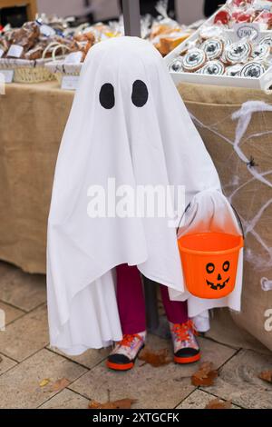 Un bambino vestito con un costume fantasma che tiene in mano un secchio di zucca arancione, davanti a una mostra di dolcetti a tema Halloween Foto Stock