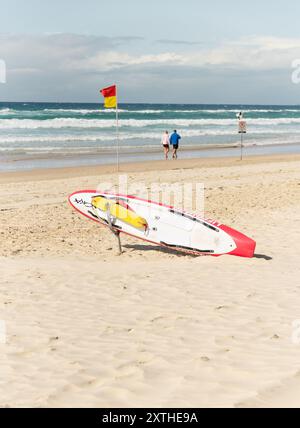 Coppia di anziani che cammina sul bordo delle acque di Surfers Paradise, Queensland, Australia. 11 agosto 2024, con equipaggiamento bagnini e tavola da surf. Foto Stock