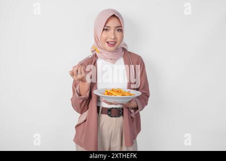 Giovane donna asiatica musulmana affamata in hijab che tiene forchetta e piatto mangiando patatine fritte su sfondo bianco isolato. Foto Stock