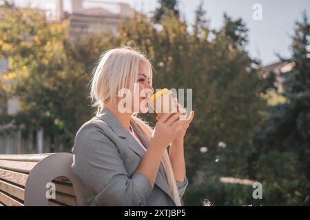 Una donna bionda siede su una panchina bevendo caffè da una tazza gialla. Indossa una giacca grigia e ha i capelli in una coda di cavallo. La scena è tranquilla Foto Stock