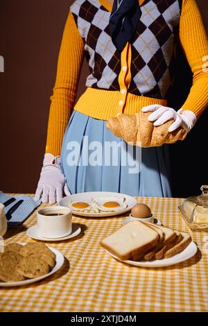 Una giovane donna elegante offre croissant al tavolo della colazione, con uova, pane, caffè. Foto Stock