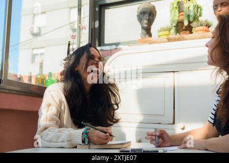 La giovane donna latina seduta a ridere ad alta voce mentre studia con la sua amica a casa, ha i capelli lunghi e sciolti, indossa un maglione bianco, ha una matita Foto Stock