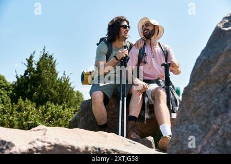 Una coppia gay gode di una giornata estiva soleggiata facendo escursioni nella natura selvaggia, condividendo una risata mentre si riposa su una roccia. Foto Stock