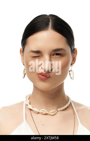 Una giovane donna con i capelli scuri strizza allegramente mentre mostra una splendida collana con conchiglie e orecchini su sfondo bianco. Foto Stock