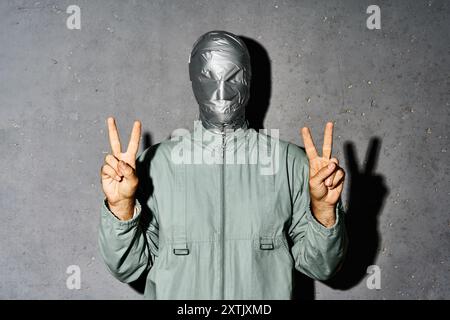 Uomo misterioso con nastro adesivo che mostra il gesto della mano con un segno V per la vittoria o la pace su sfondo grigio Foto Stock