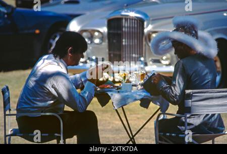 ROYAL ASCOT LADIES DAY retro 1990 elegante coppia di lusso che si gode un picnic nel terreno dell'ippodromo Royal Ascot, con la loro auto Bentley sullo sfondo. Ippodromo Royal Ascot, Berkshire. Foto Stock