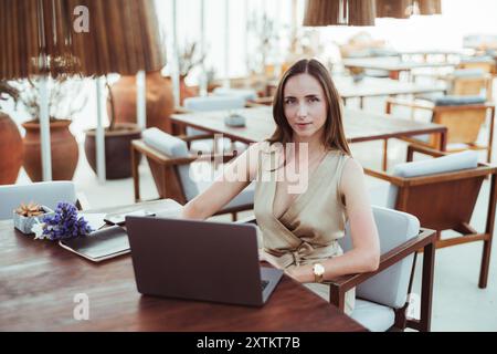 Una donna ispanica professionista imprenditrice siede su un tavolo di legno con un computer portatile in uno spazio elegante e moderno con grandi pentole in terracotta e mobili in legno Foto Stock