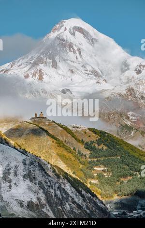 Gergeti Trinity Church, arroccata su una collina con il Monte Kazbek innevato che torreggia sullo sfondo, sotto un vibrante cielo blu in Georgia. Foto Stock