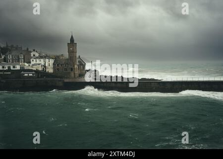 Nuvole di tempesta scure con venti di forza e onde che si infrangono contro le mura del porto e la chiesa di Porthleven sulla penisola di Lizard in Cornovaglia Foto Stock