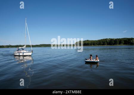 Zwei Jungs sitzen mit Paddeln auf einer Luftmatratze und treiben um die Mittagszeit entspannt auf dem Rheinsberger SEE nel Meclemburgo-Vorpommern. Ein Segelboot gleitet sanft über das Wasser. Die Jungs genießen Die Ruhe des SEE und das sanfte Schaukeln der Matratze.  7R32945 *** due ragazzi si siedono con le pagaie su un materasso ad aria e galleggiano rilassati sul lago Rheinsberg nel Meclemburgo-Pomerania occidentale a pranzo Una barca a vela scivola dolcemente sull'acqua i ragazzi si godono la calma del lago e il dolce dondolo del materasso 7R32945 Foto Stock