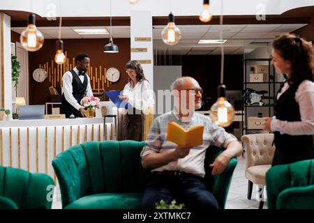 Le donne anziane firmano i documenti per il check-in alla reception dell'hotel. Il cordiale concierge maschile assiste la vecchia donna mentre compila i moduli di prenotazione, mentre la cameriera caucasica aiuta il vecchio nella lounge. Foto Stock