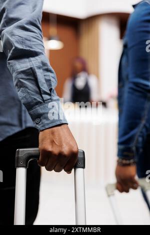 Primo piano di turisti che tirano bagagli in arrivo all'hotel, coppia afroamericana che cammina con i bagagli attraverso la hall fino al banco della reception, viaggiando insieme. Valigia per mano maschio Foto Stock