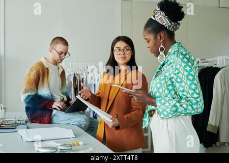 Giovane e fiduciosa designer bruna che mostra alcuni nuovi schizzi alla collega afroamericana mentre entrambi si trovano accanto al posto di lavoro Foto Stock