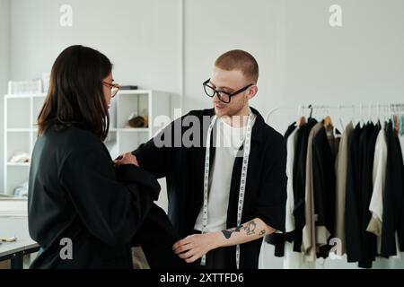 Giovane sarto maschile di occhiali da vista e abbigliamento casual davanti a una cliente che prova un blazer nero infinito nell'atelier Foto Stock