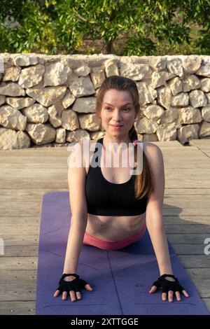 Donna che pratica yoga su un ponte di legno, indossa abbigliamento atletico rosa e nero, in un ambiente all'aperto con un muro di pietra e vegetazione sullo sfondo. Foto Stock