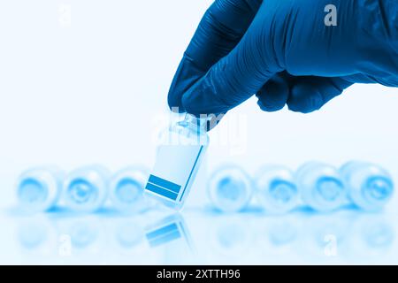 Mano del medico con i guanti blu che tiene una fiala di vaccino davanti a una fila sfocata di fiale di vaccino su sfondo blu con riflesso. Foto Stock