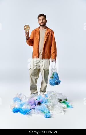 L'uomo sta in piedi su un pacco di rifiuti di plastica, tenendo una lampadina in una mano e un sacchetto di rete nell'altra. Foto Stock