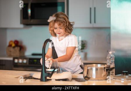 Divertenti ragazzi gemelli che aiutano in cucina con piatti di lavaggio. Bambini che si divertono con i lavori di casa. Un ragazzo carino lavare i piatti vicino al lavandino in cucina. Foto Stock