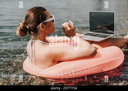 Una donna è seduta su una zattera gonfiabile rosa in acqua, usando un portatile. Concetto di relax e svago, mentre la donna si sta godendo il suo tempo al Foto Stock