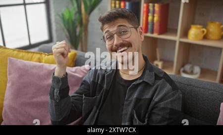 Uomo ispanico sorridente negli interni di casa, pollice rivolto all'indietro con stile casual, occhiali e baffi. Foto Stock