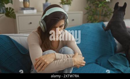 Una giovane donna ispanica siede in un accogliente salotto con le cuffie, mentre il suo bulldog francese si trova vicino su un divano blu. Foto Stock