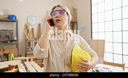 Una giovane donna con gli occhiali di sicurezza parla al telefono, tenendo in mano un casco giallo in un laboratorio di falegnameria. Foto Stock