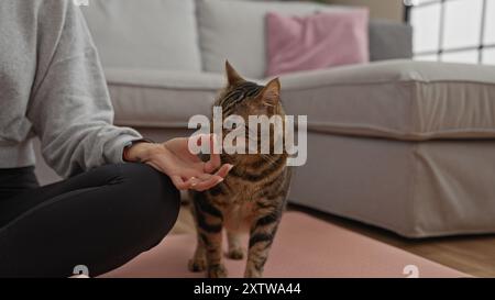 Una donna ispanica nel salotto di casa tocca delicatamente un gatto da tabby mentre si siede sul pavimento in casa. Foto Stock