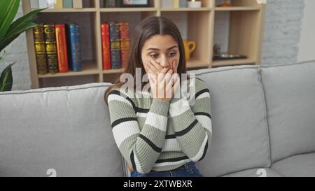 Donna ispanica seduta scioccata sul divano in casa che copre la bocca con le mani Foto Stock