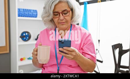 Donna dai capelli grigi in scrub controlla il telefono con la tazza di caffè in clinica. Foto Stock