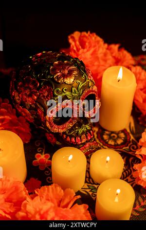 Dall'alto, un teschio decorato con fiori di metallo si trova tra le calendule arancioni e le candele accese. Lo sfondo nero e il tableclot del giorno dei morti Foto Stock