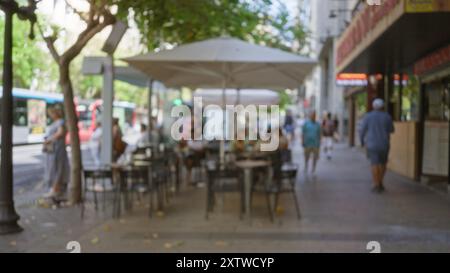 La scena dei caffè all'aperto è sfocata, con persone sedute sotto gli ombrelloni e camminate su una strada soleggiata della città. Foto Stock