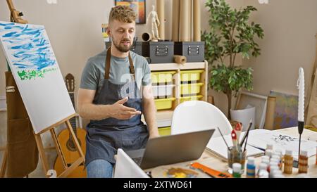 Uomo barbuto che fa una videochiamata nello studio di un artista con una tela, un dipinto e una chitarra. Foto Stock