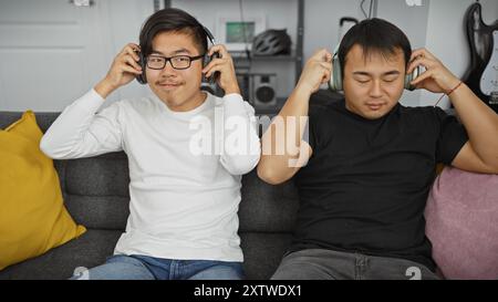 Due uomini asiatici che ascoltano musica con le cuffie in un accogliente salotto. Foto Stock