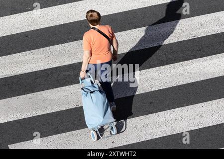 Donna anziana irriconoscibile che tira un carrello della spesa lungo un incrocio in una giornata di sole. Vista ad angolo alto Foto Stock