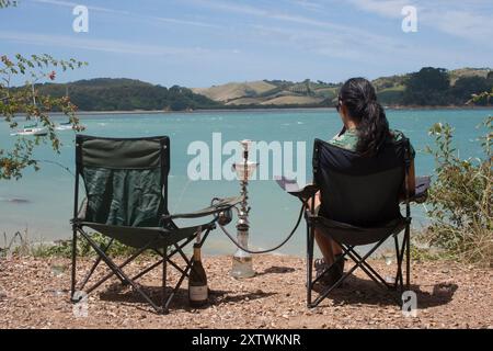 Una donna si rilassa davanti a una tranquilla vista sul lago, seduto su una sedia portatile con disposizione a narghilè e una bottiglia di vino Foto Stock