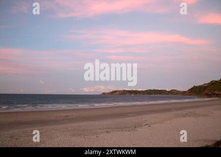 Le sfumature del sole proiettano un caldo bagliore su una spiaggia tranquilla con sabbia bianca soffice e onde calme sotto un cielo color pastello. Foto Stock